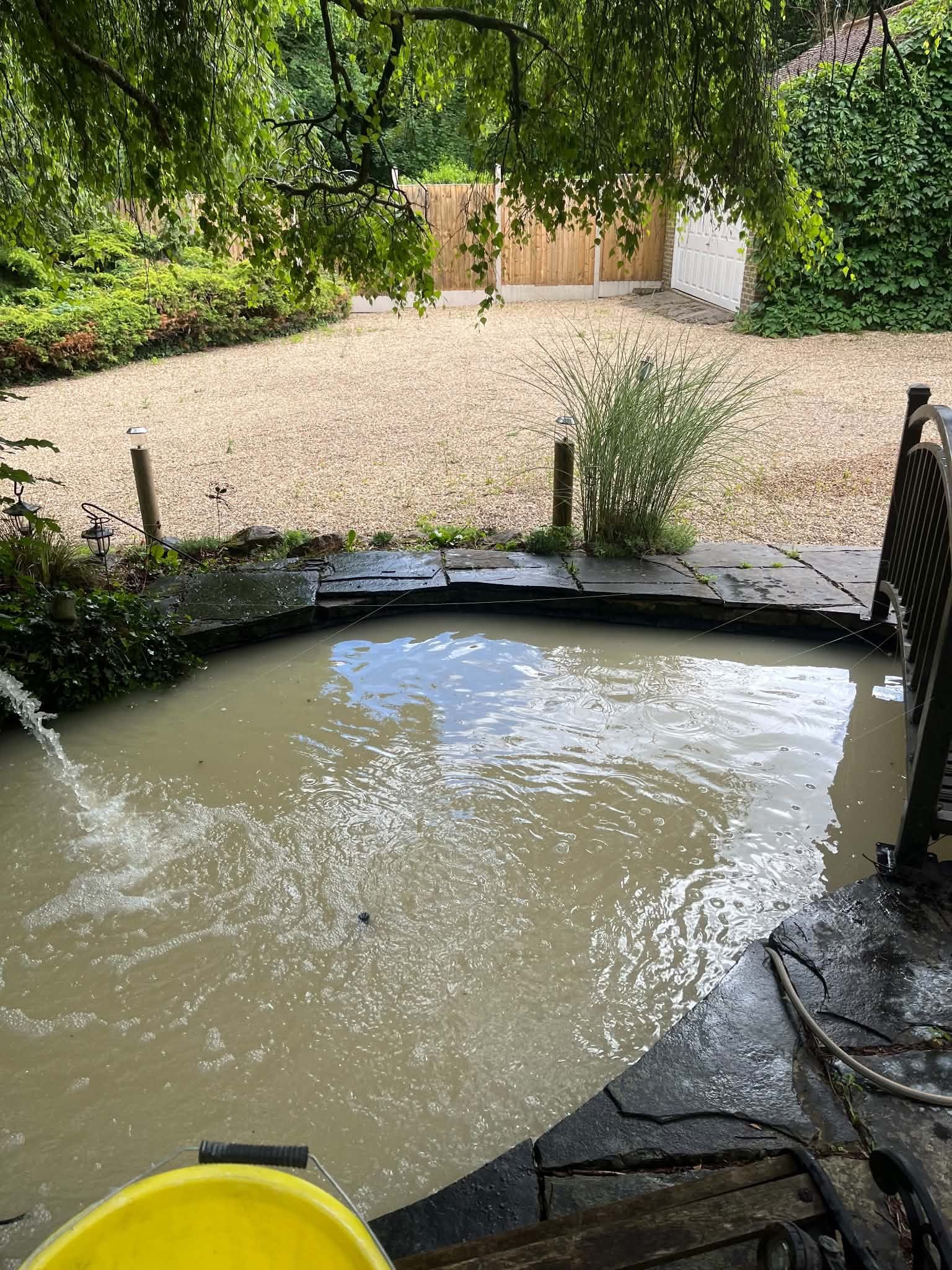A slate-edged garden pond with water entering at the left, a yellow bucket in the foreground and a bridge rail on the right
